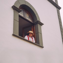 En la ventana de una parroquia con la pared blanca y lisa, una señora se asoma para ver la procesión de gente en la romería, vestida con ropa tradicional canaria.